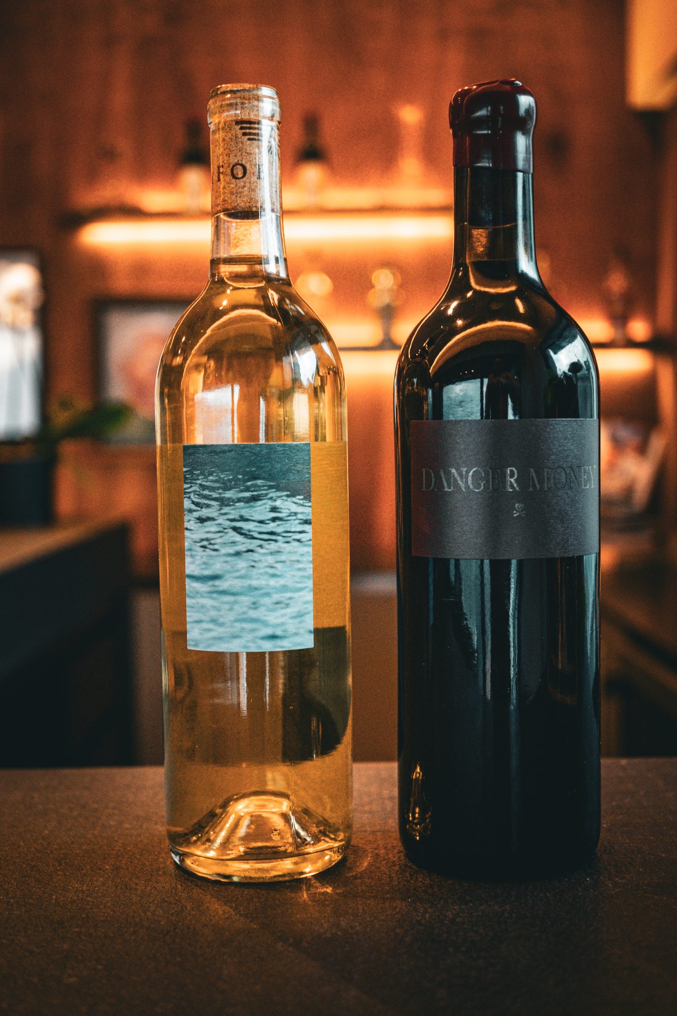 Two wine bottles on a dark counter, a white with a blue water label and a red with a black Danger Money label, in front of a warmly lit wood bar.