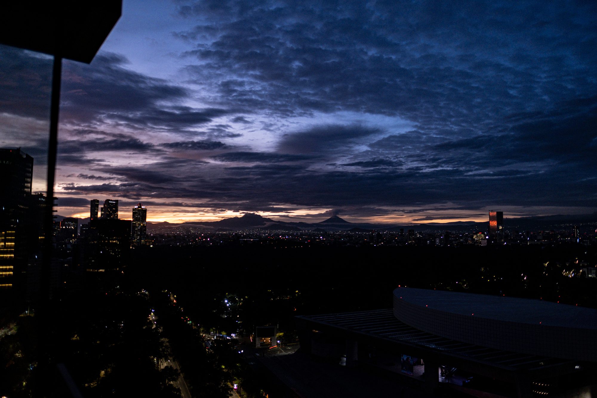 Mexico City before sunrise from a high window: dark blue clouds, an orange glow over the volcanoes on the horizon, and the city lights below.