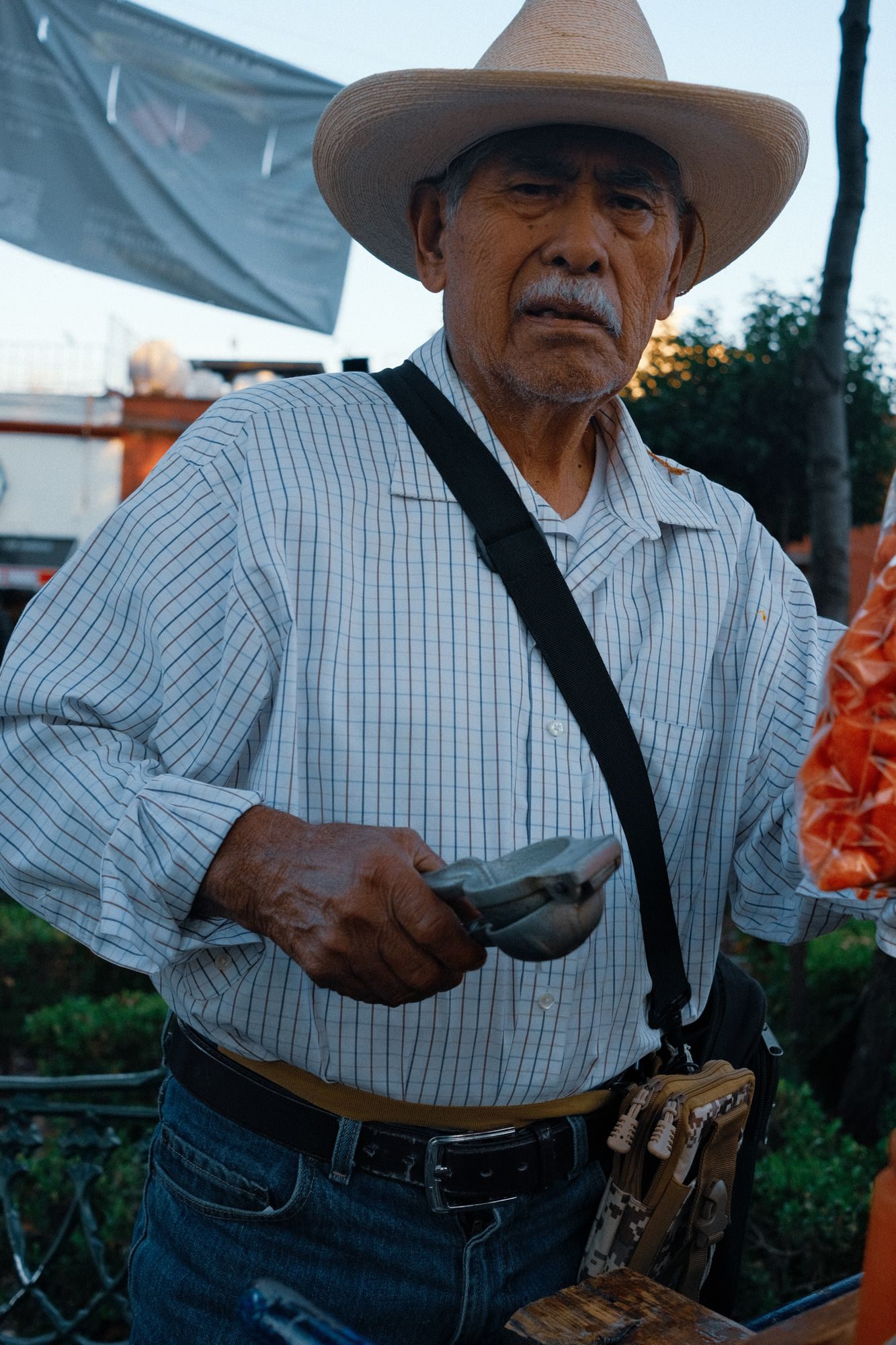 Older man in a straw cowboy hat and checked shirt holding a metal lime press and looking into the camera.