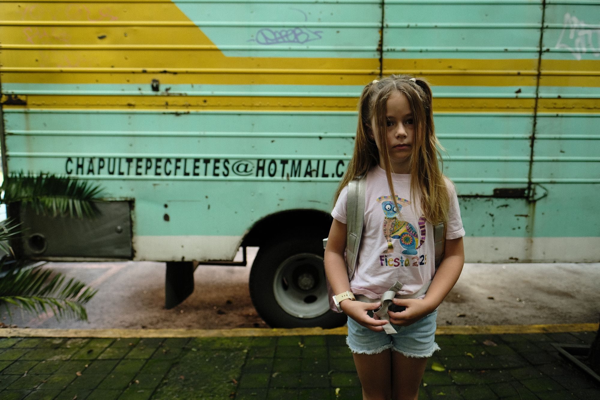 Young girl with long hair and a backpack standing in front of a weathered teal and yellow moving truck.